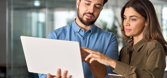 dos profesores realizando trabajo colaborativo entre docentes, uno de ellos sostiene un computador en sus manos mientras la profesora señala la pantalla